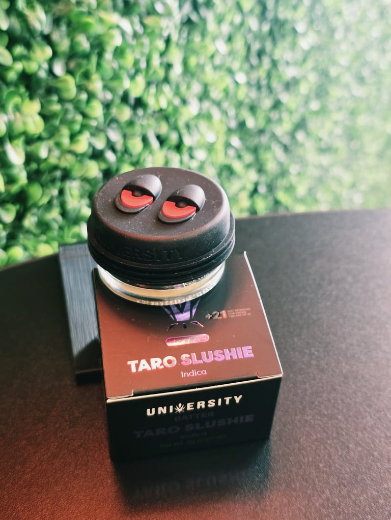 Black jar with red heart-shaped eyes labeled Taro Slushie on brown box with blurred green leafy background