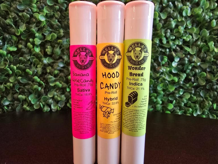 Three colorful cannabis pre-roll tubes in pink, orange, and yellow against a green leafy background