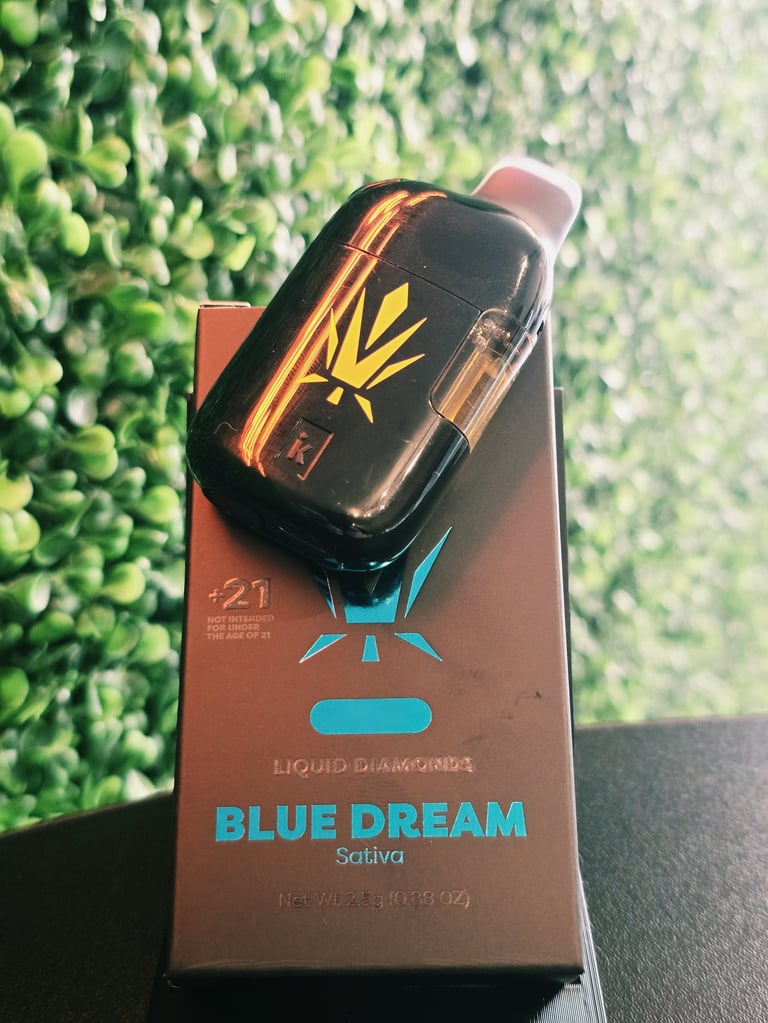Black and gold cannabis vape pen on Blue Dream sativa product box with green foliage in background