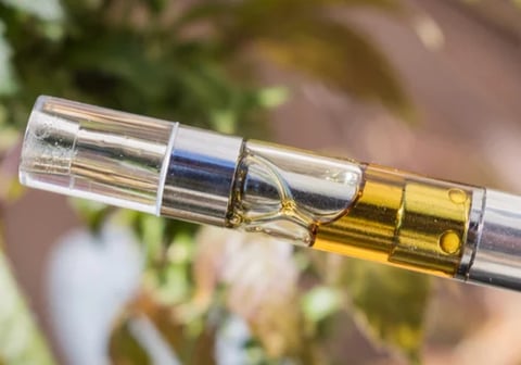 Close-up of a vape cartridge with golden liquid and clear glass chamber against a blurred green and brown background