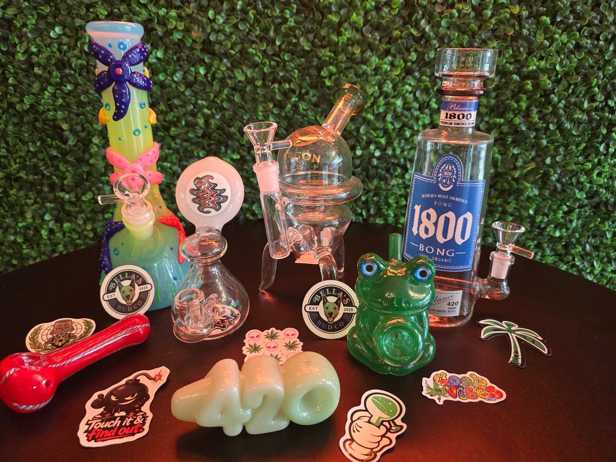 Collection of colorful glass smoking devices, decorative cannabis accessories, and 1800 tequila bottle on dark table with green ivy backdrop